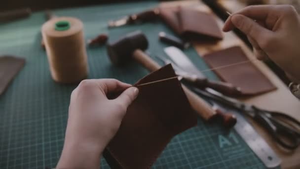 Nahaufnahme professioneller Handwerkerhände beim Nähen einer handgefertigten Ledertasche auf einem Werkstatttisch voller Werkzeuge. — Stockvideo