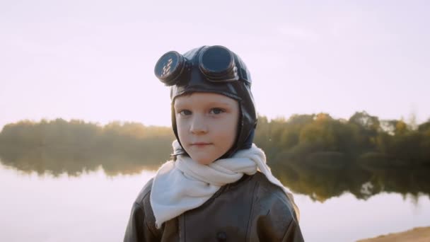Маленький мальчик смотрит в камеру, в ретро-пилотных очках и улыбается, мечтая о будущем возле закатного озера — стоковое видео
