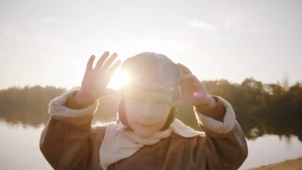 Маленький мальчик, смотрящий в камеру, в старых пилотных очках и улыбающийся в удивительном солнечном свете блики объектива близ озера замедленного движения — стоковое видео