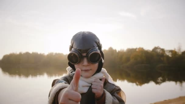 Doordachte vlieger jongetje kijken camera, een pilot bril en duimen opdagen op zonsondergang lake slow motion. — Stockvideo