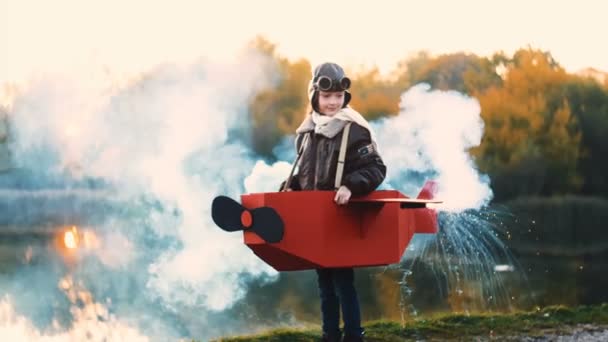 Happy little aviator girl running along sunset lake in fun cardboard plane with color smoke playing a pilot slow motion — Video Stock