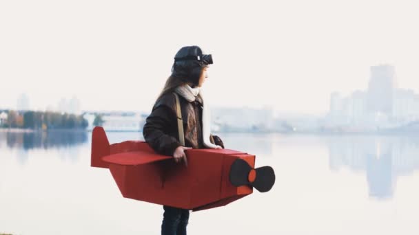 Mutlu küçük havacı kız gün batımı göl şehir panorama pilot yavaş oynamaya karton uçak kostüm, ayakta. — Stok video