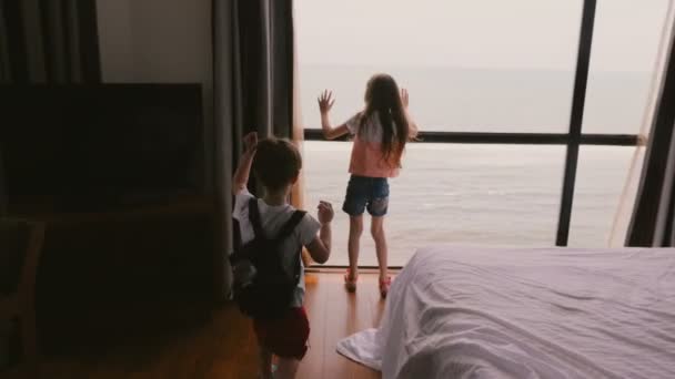 Dos niños felices emocionados corriendo hasta la gran ventana del apartamento del hotel para disfrutar de increíbles vistas al mar nublado cámara lenta — Vídeo de stock