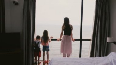 Arkadan Görünüm genç anne ve iki mutlu çocuk stand birlikte deniz görünümü yavaş hareket bakan geniş otel oda pencere.