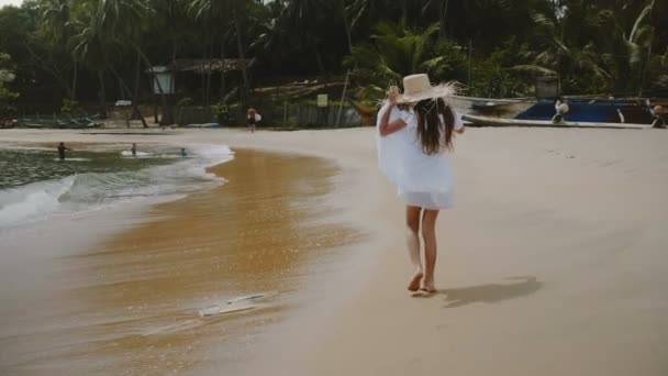 Camera follows happy excited little 5-7 years old girl walking along exotic tropical ocean beach wearing big straw hat. — Stock Video