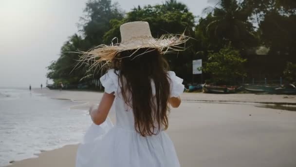 Camera follows happy carefree beautiful girl child enjoying relaxing walk along atmospheric sea beach in big straw hat. — Stock Video