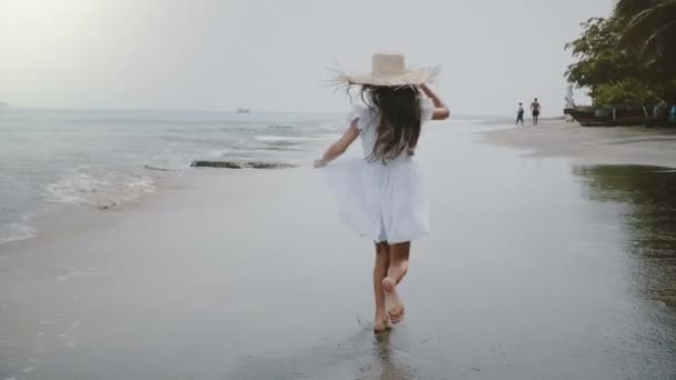 Camera follows happy beautiful little girl with long hair and straw hat running along atmospheric tropical sea beach. — Stock Video