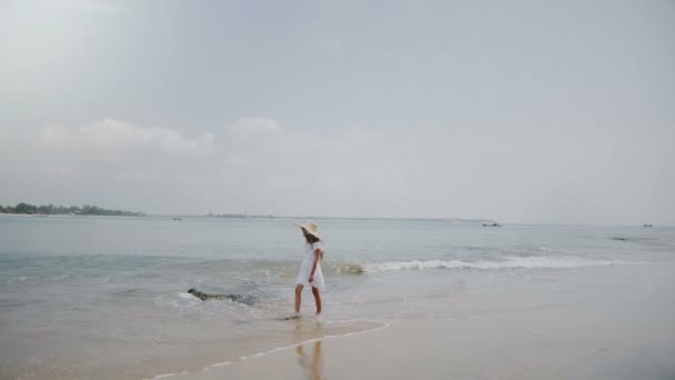 Atmospheric background shot of happy little young girl walking on waves at exotic ocean beach wearing big straw hat. — Stock Video