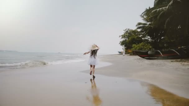 Camera follows happy beautiful little 5-7 year old girl in big straw hat running along atmospheric tropical ocean beach. — Stock Video