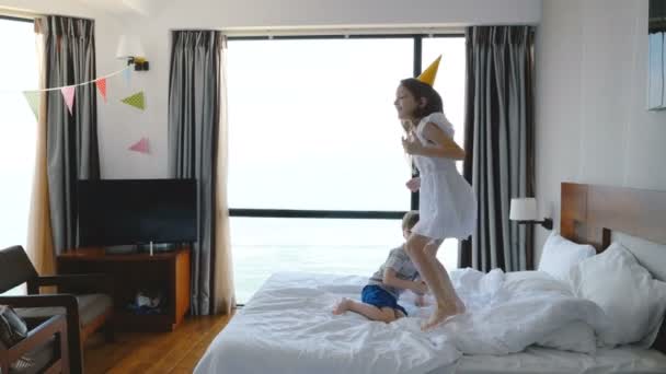 Dos niños caucásicos felices de 5-8 años, hermano y hermana, saltando alegremente en la cama en una sala de luz, divirtiéndose . — Vídeo de stock