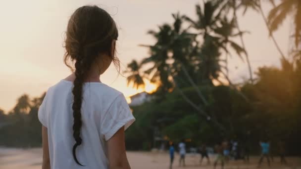Hermosa vista trasera de la pequeña niña de 6-8 años feliz en la playa tropical exótica del mar viendo la puesta de sol y la gente . — Vídeo de stock
