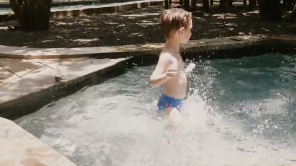 Caméra se déplace autour heureux petit 4-6 ans garçon caucasien avoir du plaisir dans une piscine d'été ensoleillée éclaboussant l'eau avec les bras . — Video