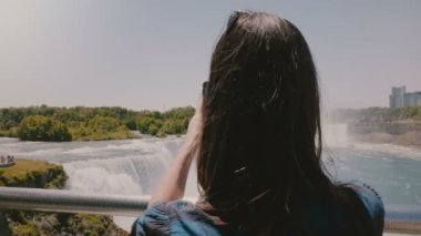 Mutlu genç kadın gezgin akıllı telefon ile epik Niagara Şelalesi Fotoğraf çekerken güzel yavaş hareket geri görünümü.