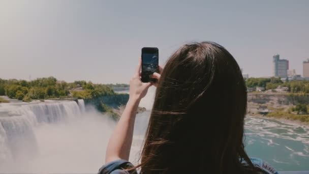 Beautiful slow motion back view shot of happy young tourist woman taking photos of incredible Niagara waterfall panorama — Stock Video