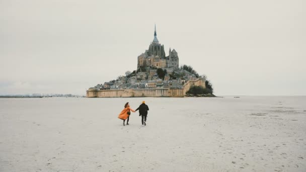 Drone камери наступним молода щаслива пара тримаючись за руки, що біжить до епічних Mont Saint Michel фортеця острів у Франції. — стокове відео
