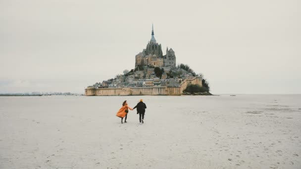 Drone kamera seuraa nuori onnellinen pari kädestä juoksee kohti eeppinen Mont Saint Michel linnoitus saari Ranskassa . — kuvapankkivideo