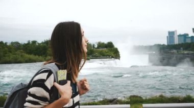 Sırt çantası, akıllı telefon ve harita boyunca yürürken ve epik Niagara Şelalesi yavaş hareket izlerken mutlu turist kadın.