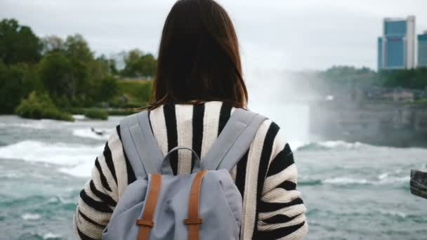Back view of happy young tourist woman with backpack watching rushing water at Niagara Falls waterfall slow motion. — Stock Video