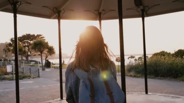 Camera follows happy casual young woman with backpack, hair blowing in the wind walking along summer sunset seaside park — Stockvideo