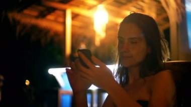 Rahat genç iş kadını yaz gecesi lounge beach cafe gece hayatı zevk akıllı telefon e-ticaret uygulaması kullanarak gülümsüyor.