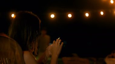 Orta atış rahat genç kadın dize ışıkları ile atmosferik yaz gecesi lounge plaj kafede akıllı telefon uygulaması kullanarak.