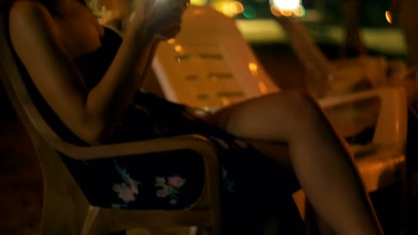 Felice giovane donna si siede in sedia da bar sulla spiaggia lounge notte, sorride utilizzando smartphone shopping online app in vacanza . — Video Stock