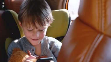Mutlu heyecanlı küçük 4-6 yaşındaki Kafkas çocuk gülümseyen, bir oyuncak ile araba çocuk güvenlik koltuğunda oturan akıllı telefon kullanarak.