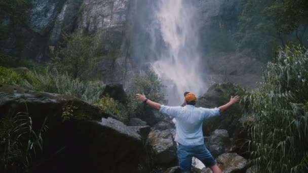 Back view excited young tourist man with arms wide open enjoying epic moments of freedom at Sri Lanka jungle waterfall. — Stock Video