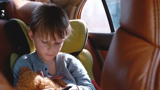 Feliz lindo niño usando la aplicación de entretenimiento de teléfonos inteligentes en el asiento de seguridad del niño del coche, mirando por la ventana en el día soleado . — Vídeo de stock