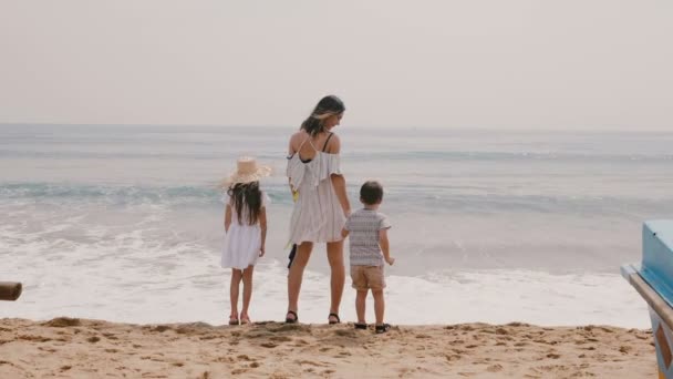 Achteraanzicht gelukkige jonge moeder met kleine jongen en meisje kijken naar de golven op vakantie in Tropical Sea Beach Slow Motion. — Stockvideo