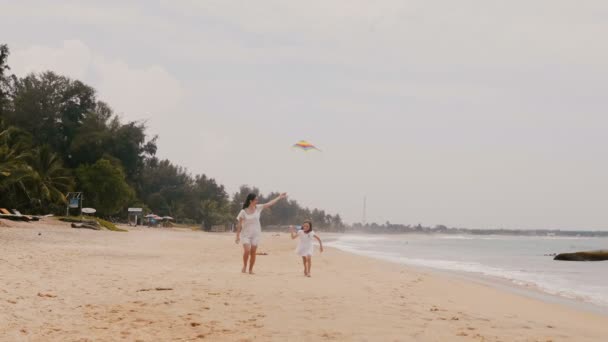 Счастливая маленькая девочка запускает воздушного змея, бежит к камере с молодой матерью на экзотический пляж тропических каникул замедленной съемки . — стоковое видео