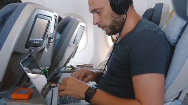 飛行機の飛行中にモバイルオフィスで働くためにラップトップを使用してスマートウォッチを持つ若い幸せな焦点を当てたフリーランスのビジネスマン. — ストック動画