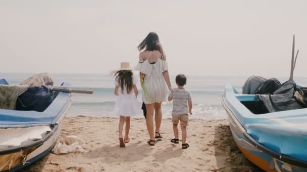 Camera follows happy young woman together with little children walking between boats towards beautiful sunny ocean beach — Stock Video