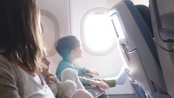 Счастливый мальчик закрывает иллюминатор самолета во время полета, глядя в камеру, улыбаясь в поездке вместе с семьей . — стоковое видео