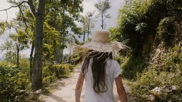 Camera follows happy little 5-7 years old girl child in big straw hat walk along sunny tropical forest road on vacation. — Stock Video