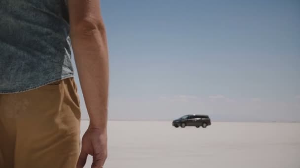 Close-up camera follows young relaxed man in casual clothes walking towards car in the middle of salt lake desert Utah. — Stock Video