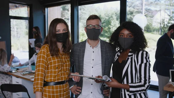 COVID-19 segurança no local de trabalho. Jovens colegas multiétnicos felizes olham para a câmera usando máscaras faciais no escritório moderno. — Fotografia de Stock