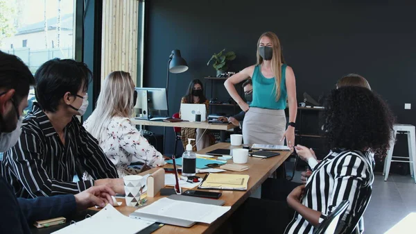 Medidas de seguridad durante la pandemia en el trabajo. Joven mujer de negocios caucásica dirigiendo reunión de oficina, empleados con máscaras. — Foto de Stock