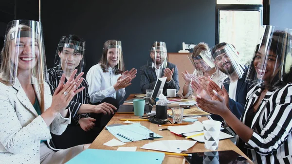 Office workplace safety measures. Happy diverse business team listening and clapping at camera, all wearing face shields
