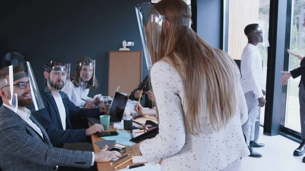 Zoom in on young happy blonde 30s female boss speaking at diverse office team meeting, all wearing plastic face shields.