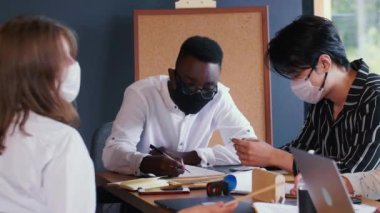 Salgın güvenliği ile iş görüşmesi. Mutlu çok ırklı iş adamları ofis masasında COVID-19 maskeleri takarak çalışıyor..