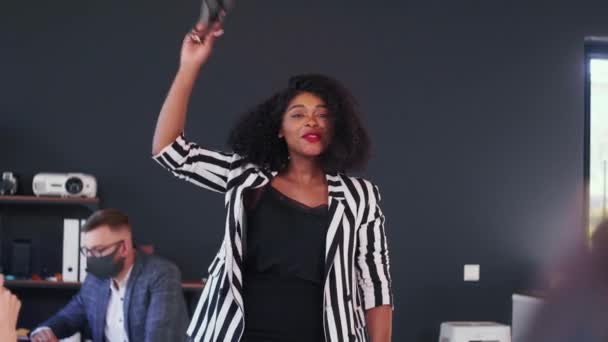 Boldog gyönyörű 20-as évek fekete női főnök veszi COVID-19 arc maszk le az irodában, csapat ünnepli együtt konfetti. — Stock videók