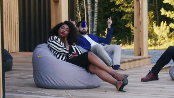 Sad birthday party. Beautiful young African woman is bored and lonely sitting on sunny terrace beanbag with friends. — Stock Video