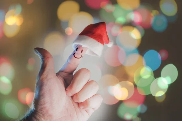 male hand with santa hat and smiley face on index finger on bokeh background