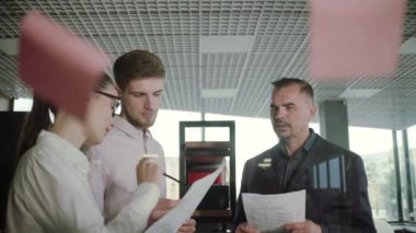 modern bir ofiste birlikte duran bir grup genç işadamı, menajerlerinin cam duvara grafik çizmesini izliyor.