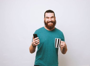 Mutlu gülümseyen sakallı adam fincan kahve ve smartphone ve kameraya bakarak holding
