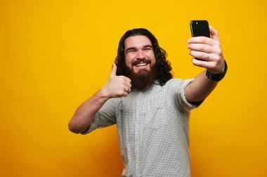 Gösterilen neşeli sakallı adam yukarı ve çekici selfie ile smartphone sarı arka plan üzerinde başparmak.