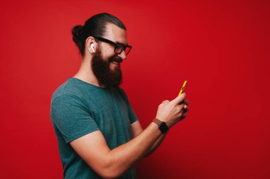 Süre ayakta ve cep telefonuyla kırmızı arka plan üzerinde izole kulaklık ile müzik dinlemek mutlu genç sakallı hippi adam portresi