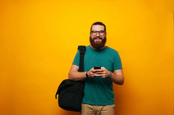 Young bearded man wearing glasses and airpods looking at the camera and smiling. Hipster wearing glasses going to work using phone.