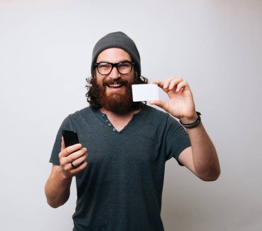 Mutlu yakışıklı hipster adam akıllı telefon ve beyaz kredi kartı banka holding ve kameraya bakarak gözlük takıyor sakallı 
